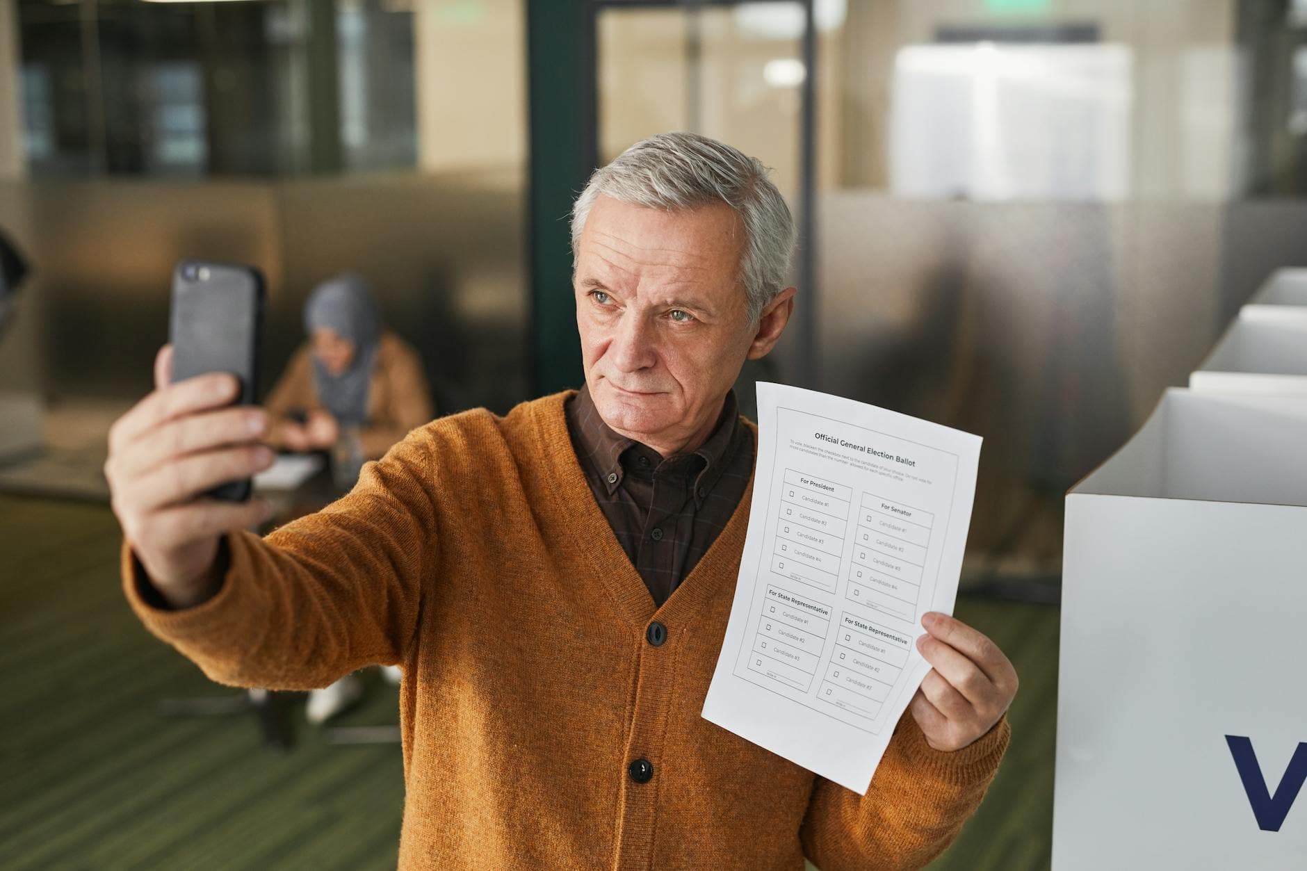 Elderly man with gray hair holding a ballot and taking a selfie in a voting booth.